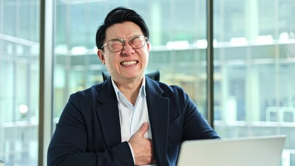 Stressed Asian businessman experiencing chest pain in modern office. Exhausted male worker having heart attack, breathing heavily at workplace. Health problems at work.