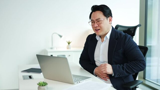 Stressed Korean businessman suffering from acute stomach pain while working on laptop in modern office. Male employee experiencing severe abdominal discomfort.