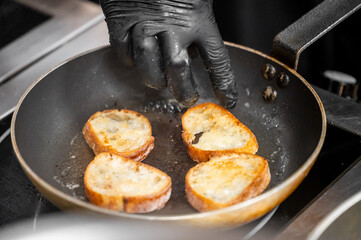 A gloved hand fries slices of bread in a pan. The golden brown bread hints at crispiness and a delicious meal in preparation, evoking a homely cooking scene.
