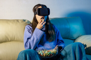 Obraz premium Young woman wearing vr headset eating popcorn on sofa at night
