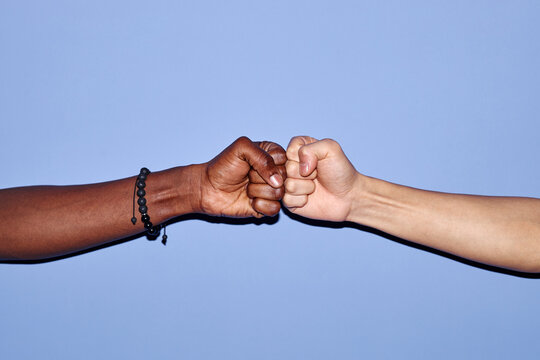Close-up of two people bumping fists against light blue background, showcasing camaraderie and solidarity. One arm dark-skinned, another light-skinned, emphasizing diversity