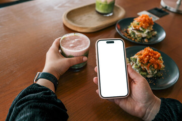 Person holding a smartphone with blank white screen and a colorful layered drink in a cafe, with...