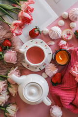 Flat lay of a cozy pink-themed setup with a gift box, flowers, candle, pink tea in a floral cup, white teapot, strawberries, and assorted sweets on a pink and fluffy background