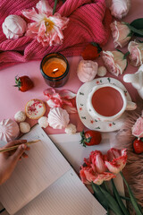 Flat lay of a cozy morning scene with pink tea, flowers, candle, meringue, strawberries, and a hand writing in a planner. Feminine and romantic setup with soft textures and warm light
