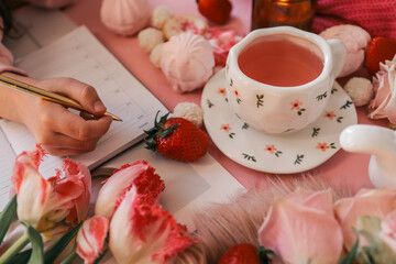Flat lay of a cozy morning scene with pink tea, flowers, candle, meringue, strawberries, and a hand writing in a planner. Feminine and romantic setup with soft textures and warm light