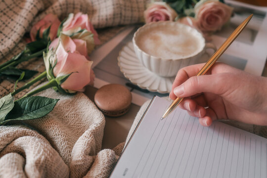 Woman writing in a notebook with a golden pen, next to a cup of cappuccino, pink roses, a macaron, and cozy fabrics. Warm, romantic flat lay for journaling or planning