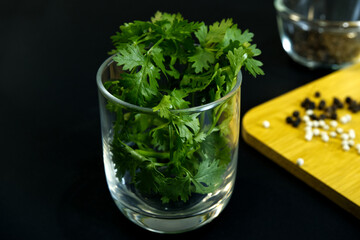 Coriander leaves in glass with blurred pepper placed on wooden board with black background. Close-up green coriander in glass. Still-life food, dry, vegetable, healthy ingredient, and plant concepts.