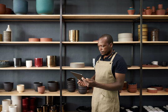 African ceramist using a digital tablet by shelves in a pottery workshop