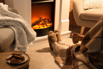 Woman in slippers and with cup of tea sitting near fireplace at home