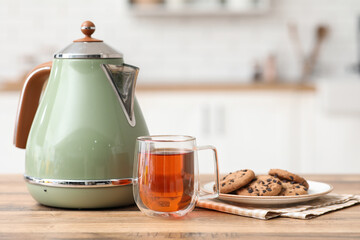 Modern electric kettle with glass cup of hot tea and sweet cookies on wooden table in kitchen