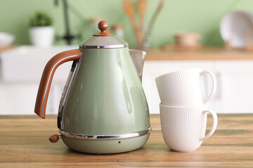 Modern green electric kettle and cups on wooden table in kitchen