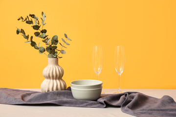 Vase with eucalyptus and dishes on tablecloth against yellow background