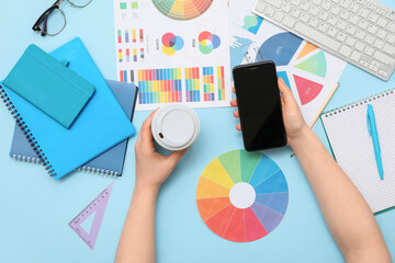 Female graphic designer with blank mobile phone, coffee and color palettes on blue background, top...