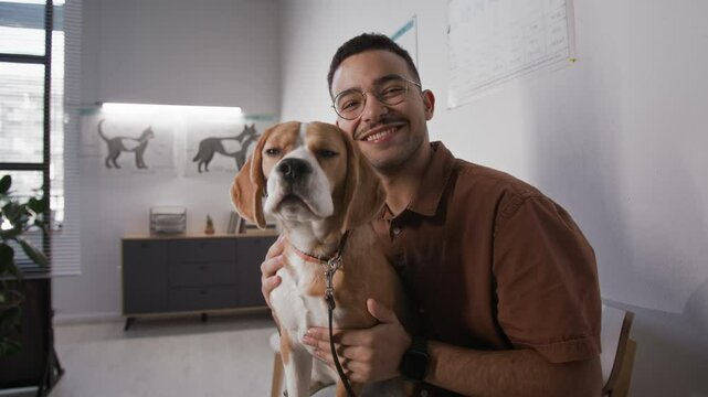 Portrait of young joyful man sitting with cute beagle in modern veterinary clinic, petting yawning dog and posing for camera with smile