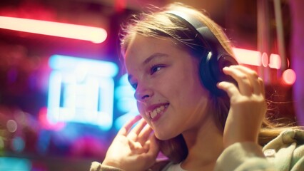 Positive Caucasian Asian teen child girl listening music headphones in city neon schoolgirl kid...