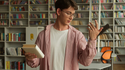 Caucasian teen man teenage male teenager guy boy student schoolboy pupil confused puzzled choose between book and mobile phone modern technology choosing reading literature education in library school