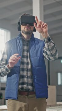 Focused Caucasian man wearing VR headset while standing in industrial environment. Male interacting with virtual interface. Testing industrial process in augmenter reality while reaching out with hand