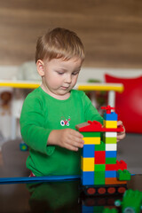 A young child focuses intently on constructing a tall tower using various colorful building blocks in a well-lit play area. The atmosphere is cheerful and playful