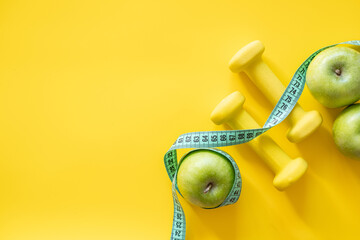 Healthy lifestyle concept with green apples, yellow dumbbells, and measuring tape on yellow...