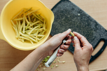 Hands are peeling green beans over a yellow bowl on a wooden table. A board is lying nearby, and...