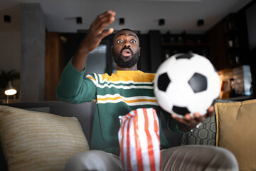 A surprised man reacts dramatically while watching a soccer match on TV at home. He sits on a couch...