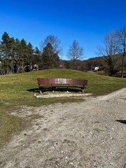 Obraz premium A rustic wooden bench sits atop a small grassy hill, surrounded by lush greenery and a gravel path. The backdrop features a clear blue sky, distant trees, and a few charming houses nestled 