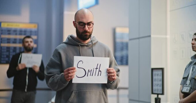Transportation Service in International Airport Terminal: Airport Greeter Holding Personalized Greeting Sign, Waiting for Passengers From Plane Flight. Diverse People, Travelers Walking. Medium Shot.
