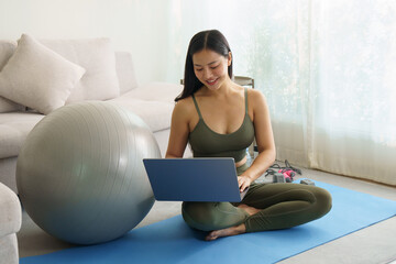  Young woman in activewear sits on a yoga mat and using laptop during home workout.
