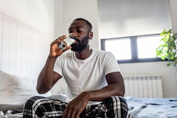 A young man sits on his bed in the morning, drinking a glass of water. He looks refreshed and relaxed, starting his day with hydration in a bright and cozy bedroom setting.