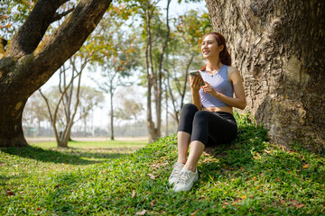 Happy young woman in athletic wears sitting under a large tree, relaxing after a workout in the park.