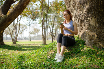 Happy young woman in athletic wears sitting under a large tree, relaxing after a workout in the park.