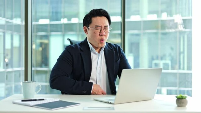 Stressed Asian businessman having a stomach ache in the office. He is sitting at his desk with his hand on his stomach, indicating pain and stress.