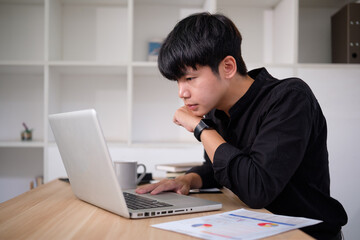 Young businessman in a black shirt intently works on his laptop, analyzing data and documents on his desk.