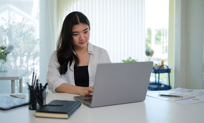 Focused woman in a white striped shirt is engaged in work on her laptop at a well-organized desk.