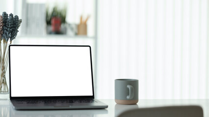 Sleek laptop with a blank white screen sits on a modern white desk, accompanied by a gray coffee mug and dried flowers in a vase.