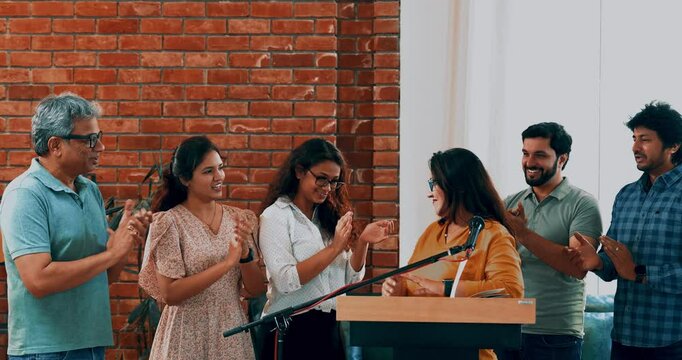 Indian Asian young entrepreneur confidently delivering speech to aspiring business professionals at conference, standing at the podium on mic as her team joins from behind, clapping and cheering