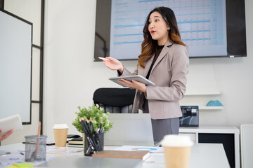 Confident woman in a beige blazer holds a tablet and gestures while leading a discussion in a corporate meeting room.