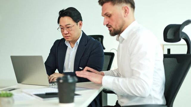 Two diverse businessmen discuss paperwork and plans while seated at a desk, utilizing a laptop. Collaborative business in a modern workspace.
