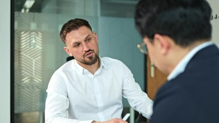 Two businessmen, one Caucasian with a beard and one Asian, sit and talk seriously during an important business meeting in a modern office.