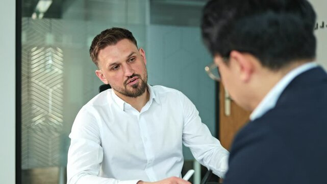 Two businessmen, one Caucasian with a beard and one Asian, sit and talk seriously during an important business meeting in a modern office.