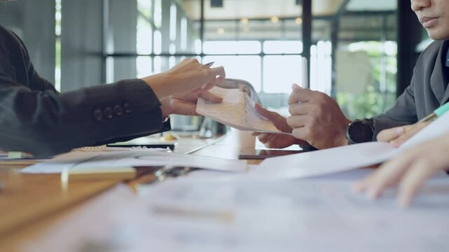 Two people are sitting at a table with papers and pens. One of them is writing on a piece of paper. Scene is focused and serious, as the two people are working on something important