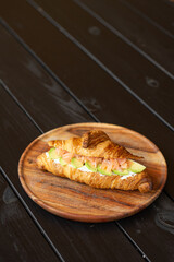 Freshly baked croissant sandwich with salmon and avocado on wooden plate, minimal rustic composition for menu or advertisement
