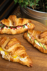 Three gourmet croissant sandwiches with different fillings on wooden board: peach and walnut, salmon and avocado, cured meat and herbs – rustic outdoor brunch