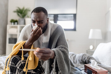 A sick man wrapped in a blanket sits on his bed, coughing into a tissue while checking his...