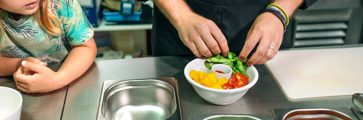 Banner of unrecognizable chef carefully preparing a vegan poke bowl while little girl watches him attentively in professional kitchen. Healthy eating and cooking education concept.