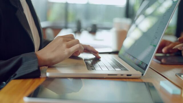 A woman is typing on a laptop in a cafe. The laptop is open and the woman's hand is on the keyboard. There are other people in the background, and a few items on the table, including a cell phone