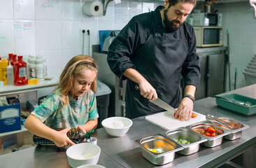 Professional chef cutting cherry tomatoes while his young daughter is pouring soy sauce oil in a...