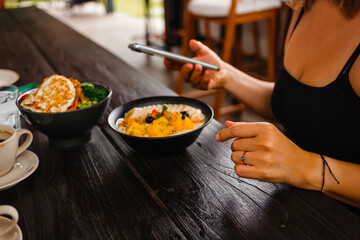 Woman has breakfast with delicious oatmeal with bananas, mangoes and berries, she takes pictures on mobile camera and posts it on social networks. Concept of counting calories using a mobile app