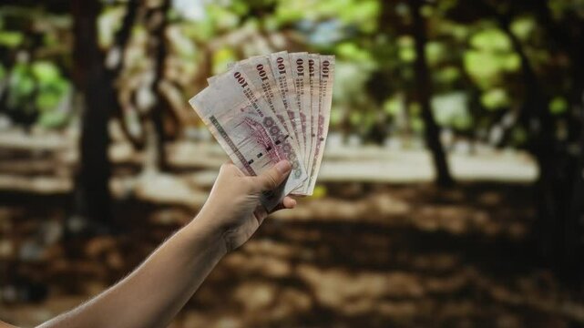 Man holding saudi riyal banknotes in hand outdoors in a park, showcasing currency against a natural garden background.