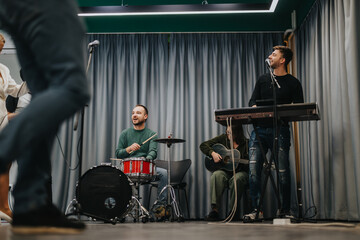 A lively band rehearsal featuring three musicians playing drums, guitar, and keyboard. The setting...